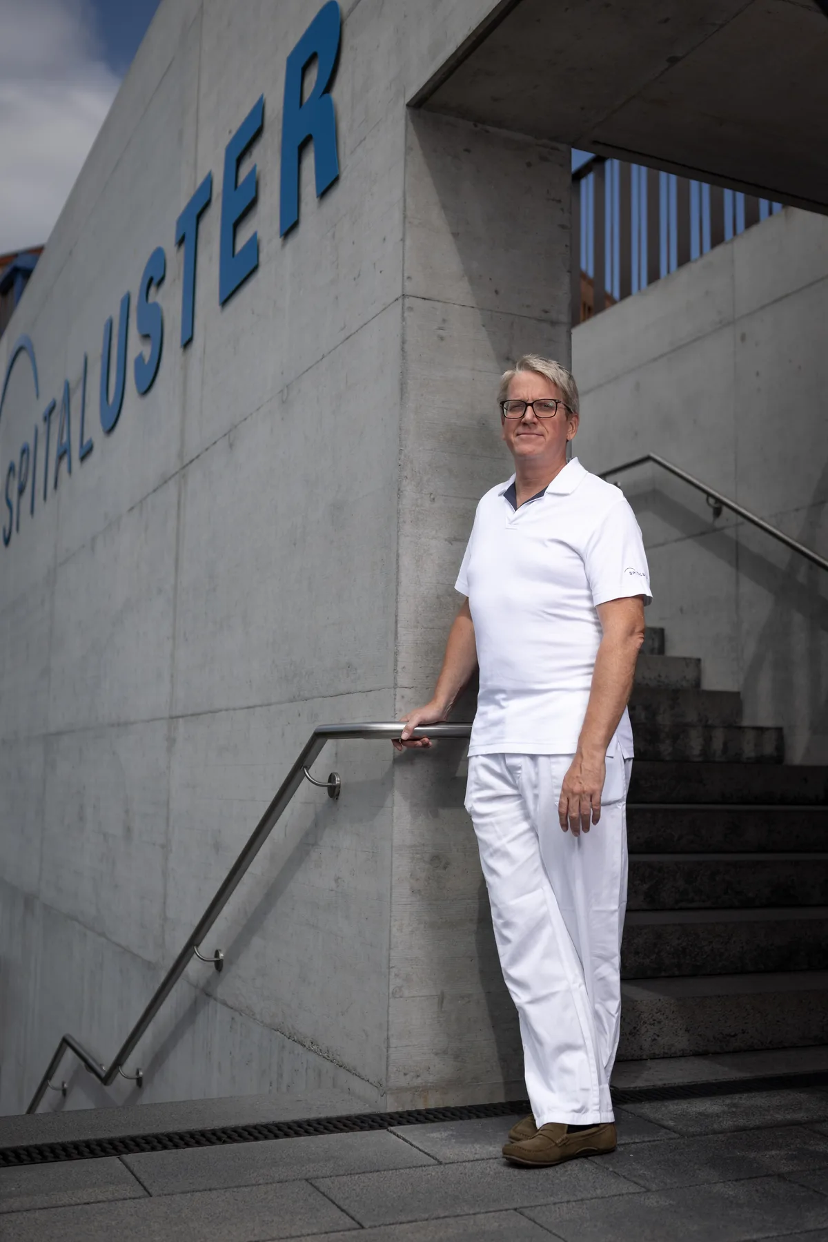 Ein Mann in weissen Spitalkleidern steht bei einer Betontreppe.